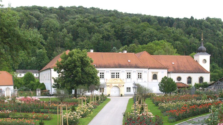 Rose Garden Pitten, © Rosengartenverein Pitten A historic building with a red roof, surrounded by a rose garden and trees, with a wooded hill in the background.