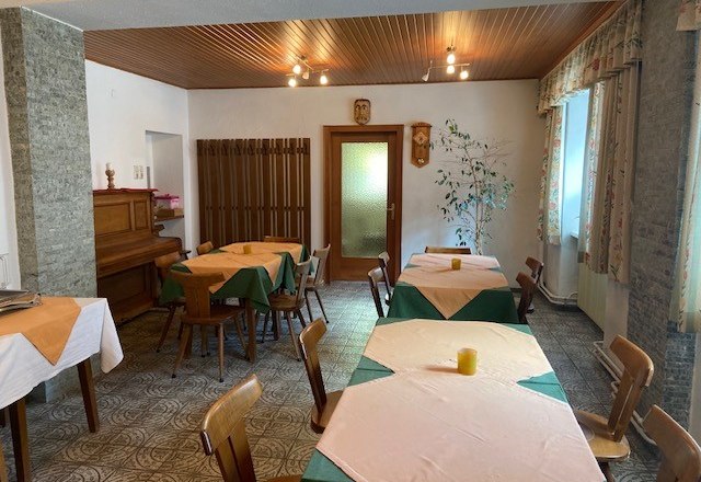 Breakfast area, © Wiener Alpen A cozy breakfast room with wooden tables, chairs and a piano. The tables are covered with tablecloths and there are windows with curtains.
