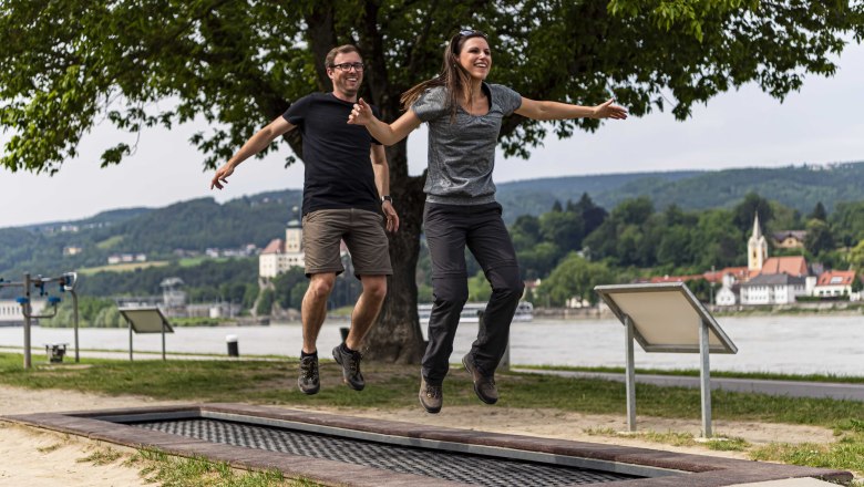 Movement park Ybbs, © Robert Herbst Two people jumping on a trampoline outdoors with a river and buildings in the background.