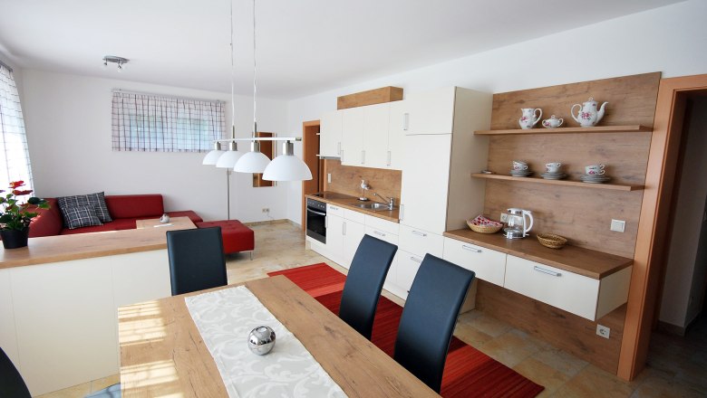 Living and dining area with kitchen, © Familie Harrauer Modern living and dining area with open kitchen, red sofa, wooden table and white hanging lamps.