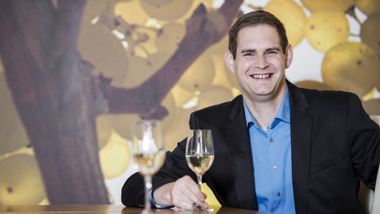 Martin Blaha, © Alfred Kusdat A man in a suit sits at a table with two wine glasses in front of a background of grapes.