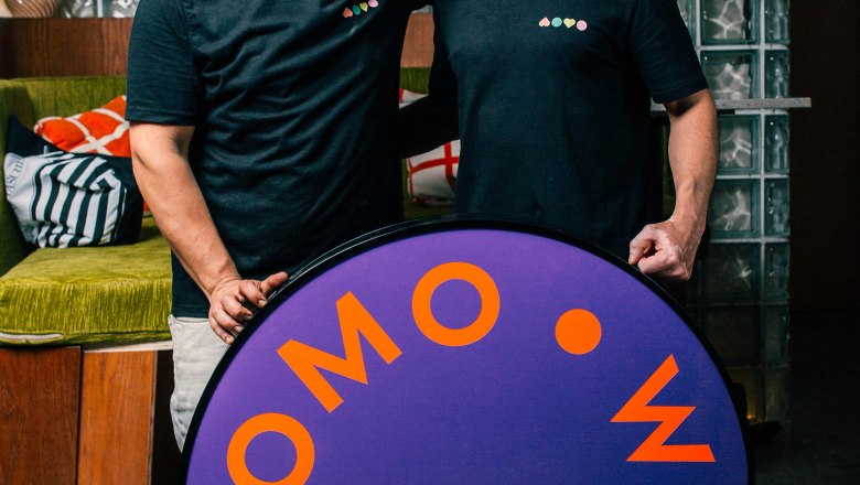 Logo is held by the owners, © Womo Two men in black T-shirts stand smiling behind a purple sign that reads 'WOMO'.