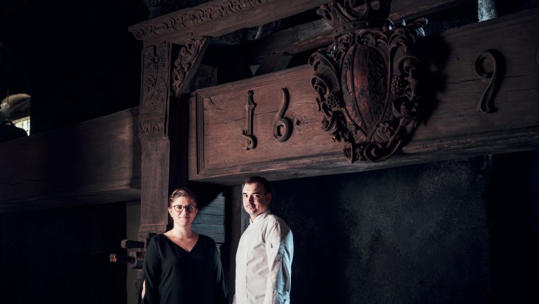 Hosts: Bernadette and Samuel Pope, © Niederösterreich Werbung/Julius Hirtzberger Two people stand in front of an old wooden beam with carvings and the date 1692.