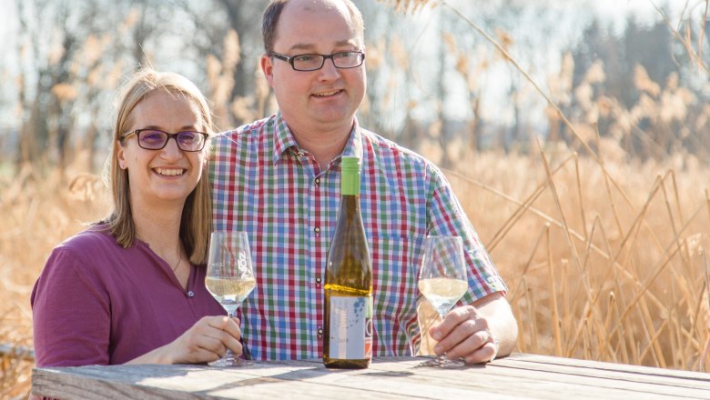 Owner family, © Verena Saloukeh Photography A man and a woman are sitting at a wooden table outside with wine glasses and a bottle of wine.