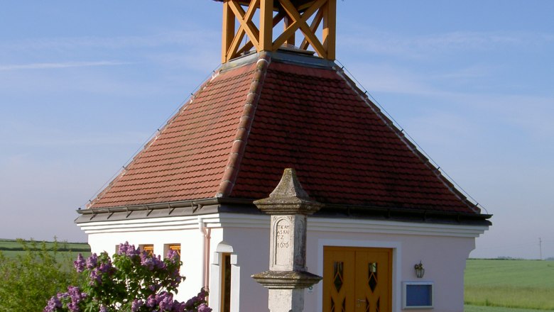 Market town of Windigsteig, © Marktgemeinde Windigsteig Small chapel with a red roof and cross, surrounded by flowers and a bench, under a clear sky.