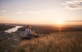 Braunsberg, VIA.CARNUNTUM., © Donau Niederösterreich, Andreas Hofer A couple sits on a hill and looks out over a river and the landscape at sunset.