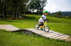 Mini Bikepark Wexl Trails, © Wexl Arena St. Corona am Wechsel A child with a green helmet rides a balance bike over a wooden ramp in the Mini Bikepark Wexl Trails.