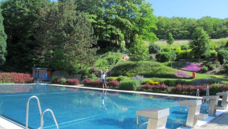 Waldbad Wöbling, © Marktgemeinde Wölbling An outdoor pool with clear water, surrounded by a green landscape and colorful flowerbeds. A person stands at the edge of the pool.