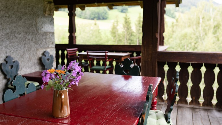 Sun veranda, © Wiener Alpen - Lierzer A rustic veranda with wooden tables and chairs, decorated with a jug full of colorful flowers. A green landscape can be seen in the background.
