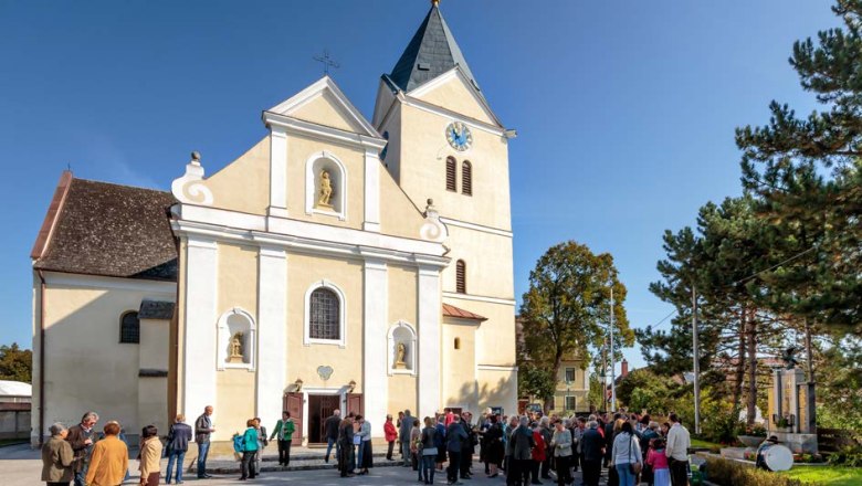 Church square, © Gemeinde Prottes People gather in front of a church with a tower and clock in sunny weather.