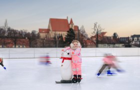 Ice rink in Eggenburg, © Martin Mathes A child in pink clothing stands on an ice rink with a penguin aid. A church can be seen in the background.