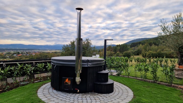 Johannesbachklamm Chalet Hot Tub, © Johannesbachklamm Chalet A round hot tub with an outdoor fireplace, surrounded by green countryside and cloudy skies.