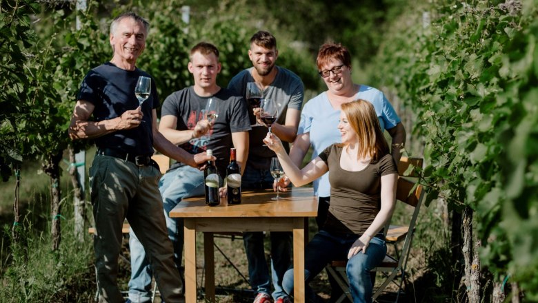 Hofer family, © Astrid Bartl A group of five people sit and stand at a table in the vineyard, holding wine glasses and smiling.