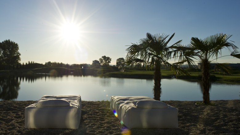 Bathing beach, © DCC Two loungers on the shore of a lake with palm trees and sunset in the background.