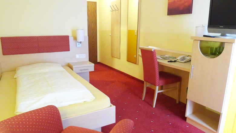 Red Rooster, © Gasthof-Pension Böck A hotel room with a single bed, red carpet, desk and chair. A picture hangs on the wall, next to it a television on a cupboard.