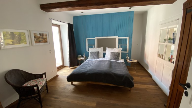 1st bedroom, © Kathrin Bürger Modern bedroom with wooden floor, blue accent on the wall, double bed and rattan chair.