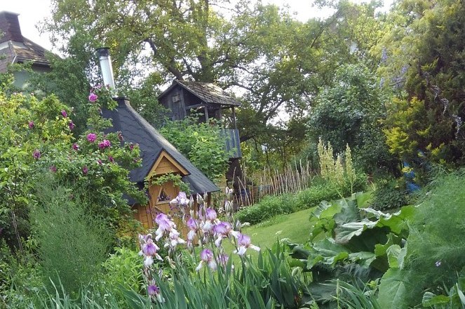 finnhaus-bluetenwald3_excerpt_c_edith-white, © Edith Weiß An idyllic garden with a small wooden house, surrounded by flowering plants and trees.