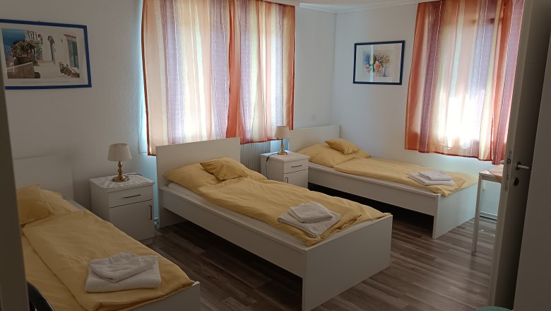 TDR-Gasthof Schwarzer Adler- Bedroom, © Márton Varga Three single beds in a bright room with yellow bed linen and colorful curtains.