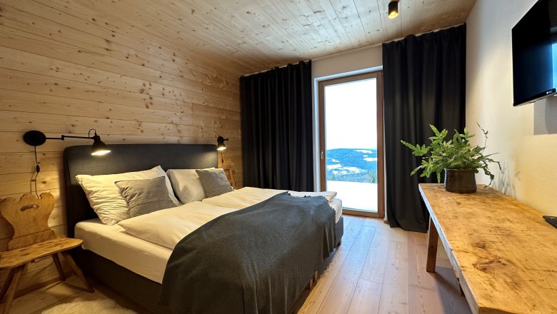 Vacation home Orthof - Bedroom, © Ferienhäuser Gruber-List Cozy bedroom with wooden walls, double bed, wooden chair, table with plant and large window with curtains.