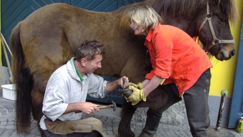 Old farriery and wagon smithy, © gutger A farrier and a woman are working on a horse's hooves in front of a blue door.