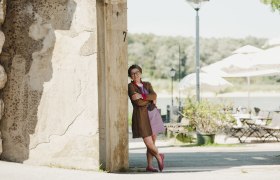 Landlady Michaela Brenner-Melchior, © Niederösterreich Werbung/Michael Reidinger Smiling woman leaning against a wall, in the background a terrace with tables, chairs and parasols.