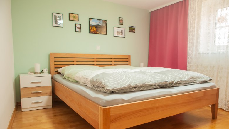 Bedroom, © Fam. Heinrich A bedroom with a wooden bed, green wall, pictures, bedside table and pink curtains.