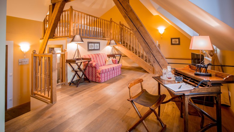 Romantic suite, © Schlossschenke Cozy attic suite with wooden beams, sofa, desk and stairs to a sleeping area.