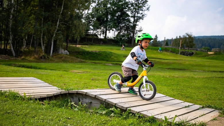Mini Bikepark Wexl Trails, © Wexl Arena St. Corona am Wechsel A child with a green helmet rides a balance bike over a wooden ramp in the Mini Bikepark Wexl Trails.