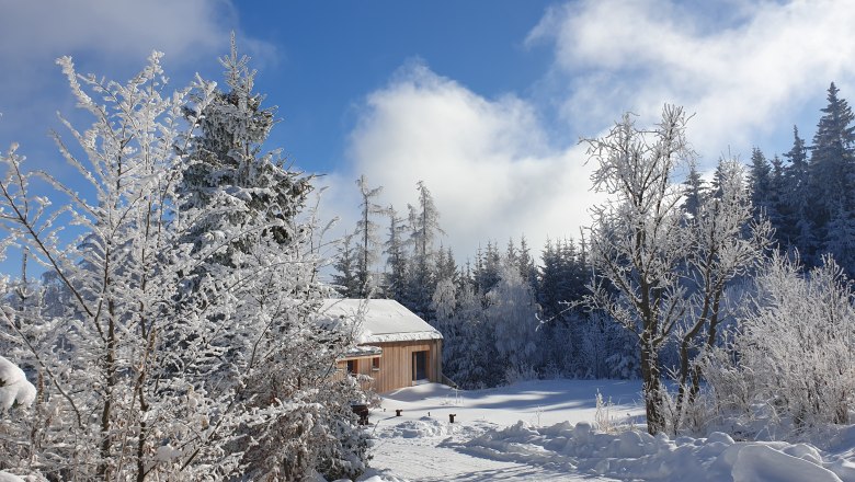 Vacation homes Gruber-List, © @Ferienhäuser Gruber-List Snowy landscape with a wooden house and trees under a blue sky.
