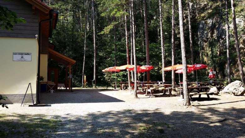 Terrace at the Eisensteinhöhle house, © Alpenverein Wiener Neustadt Terrace with tables and red parasols in the forest next to a building.