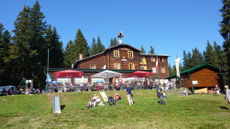 Hallerhaus, © Hallerhaus, Foto Heinz Bayer Wooden house in the mountains with people and bicycles in front of it, surrounded by trees.
