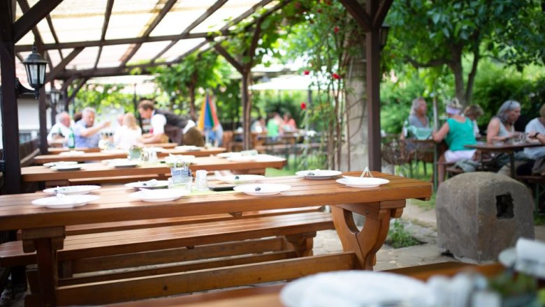 The guest garden, © Alexander Pfeffel A guest garden with wooden tables and benches, surrounded by green plants. People sit in the background and enjoy their meals.