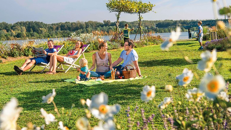 Garden City Tulln, © Stadtgemeinde Tulln/Robert Herbst Family picnic in the countryside with flowers in the foreground and a river in the background.
