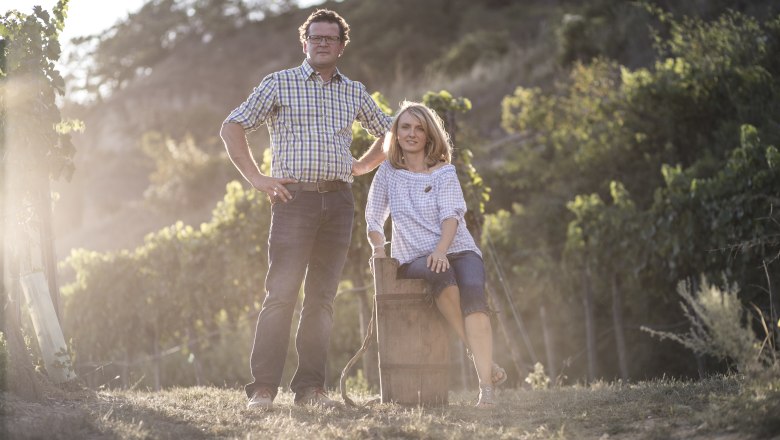 Sonja and Markus Oßberger, © Nino Zimmerhackl A man and a woman are standing in a vineyard, the man standing and the woman sitting on a barrel. The sun is shining through the leaves.