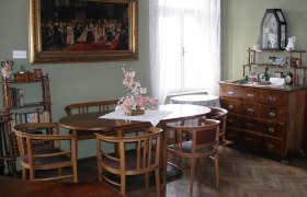 Home, © Stadtgemeinde Mank A traditionally furnished living room with wooden furniture, a round table, chairs, a chest of drawers and a large painting on the wall.