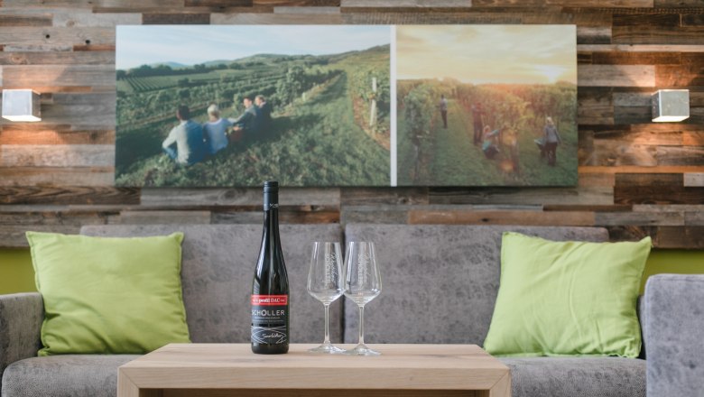 Apartment, © schwarz-koenig.at Living room with gray sofa, green cushions, wooden table, wine bottle and glasses, mural with vineyard.