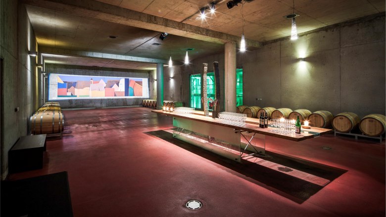 Barrique cellar, © Helge Kirchberger A modern wine cellar with wooden barrels and a long table with glasses and bottles.