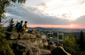Viewing platform at the Mandelstein, © Studio Kerschbaum Two people on a rock formation at sunset with a view of a viewing platform and vast landscape.