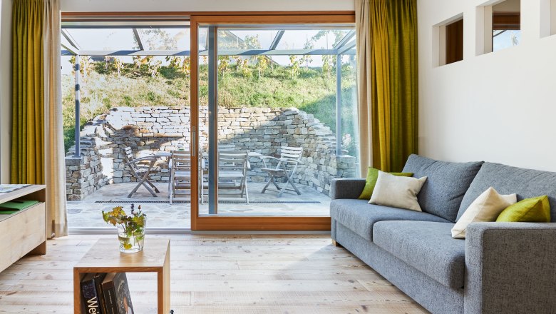 Vacation at the Hindler Winery, © Andreas Hofer Modern living room with gray sofa, wooden floor and large windows overlooking a terrace with stone wall and garden furniture.