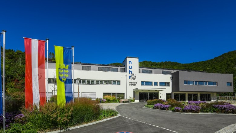 House view, © Nuhr Medical Center Modern building with three flags in front, surrounded by green landscape.