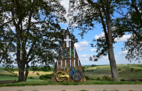 Cyclist Marterl in Mittergrabern, © Christian Häusler A shrine with a yellow and blue bicycle, surrounded by trees and fields under a blue sky.