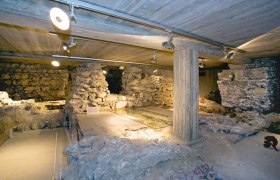 Lower church, © Stadtgemeinde Traismauer Underground ruins with stone walls and concrete pillars, illuminated by ceiling spotlights.