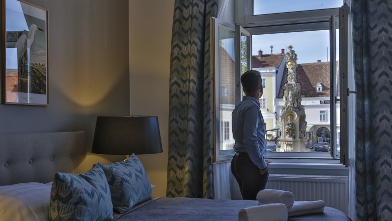 sgrafit Hotel, © Astrid Bartl Person stands at the window of a hotel room and looks at a fountain outside.