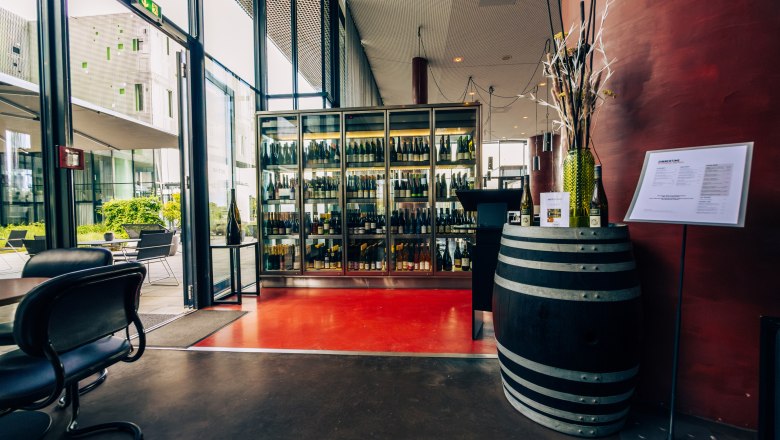 LOISIUM Langenlois, © Niederösterreich Werbung / Maximilian Pawlikowsky Interior view of a modern wine cellar with a glass cabinet full of wine bottles and a wine barrel as decoration.