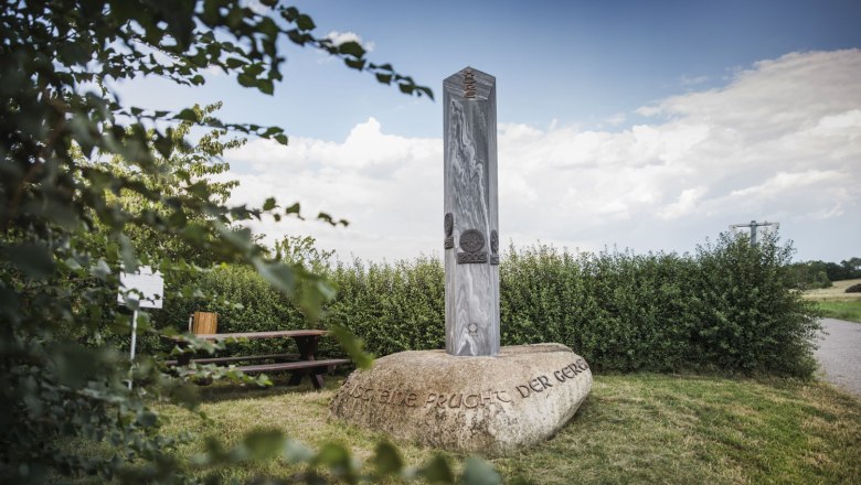 Millennium Column, © Daniela Matejschek A stone pillar stands on a large rock with an inscription, surrounded by bushes and a picnic table in the background.