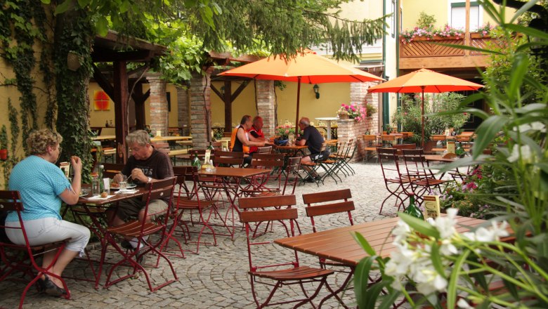 Gausterer, © Gausterer Markus A cozy beer garden with wooden tables, red chairs and orange parasols. People sit and eat outdoors.