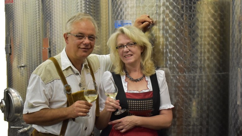 Cellar, © Weingut Schober A man and a woman in traditional dress stand in front of wine tanks and hold wine glasses.