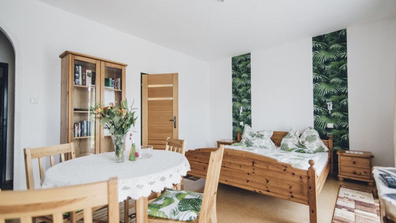 Double room, © Józef Swierkot A cozy double room with wooden furniture, a table with flowers, a bed with a plant pattern and a bookshelf.