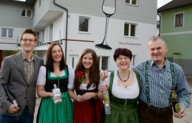 Krenn family, © Wirtshausbrennerei Krenn, Johann Frank A family stands in front of a building labeled 'Haus Peilsteinblick'. They are wearing traditional clothing and holding bottles in their hands.