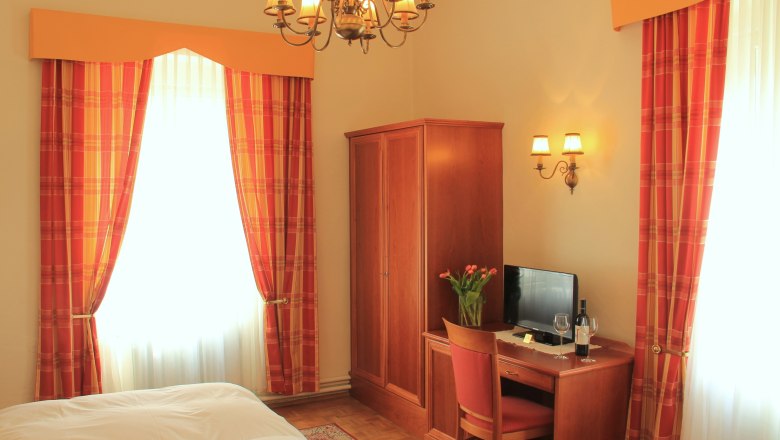 Double room Superior, © Schendl A cozy hotel room with a double bed, wooden furniture, a desk with TV and a vase of flowers. Curtains in red-orange.